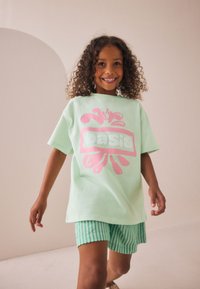 Tricou verde deschis din bumbac cu un imprimeu roz îndrăzneț pe care scrie "oasis", asortat cu pantaloni scurți în dungi albastre și albe, pe un fundal neutru.