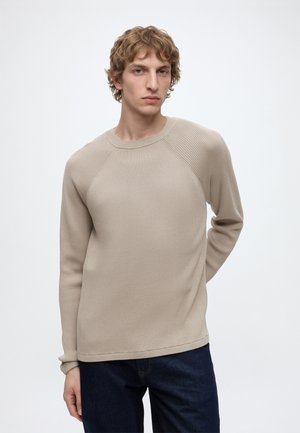 Jonge man met kort krullend haar, draagt een beige geribde trui met lange mouwen en donkere jeans, staand tegen een effen witte achtergrond.