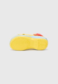 Sabot pour enfant bleu et rouge avec semelle jaune, vu par le dessous sur un fond gris uni.