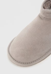Chinelo de camurça cinza com uma textura suave, bico arredondado e detalhes cosidos à volta da sola. Projetado para conforto e calor.