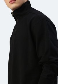 Schwarzer Reißverschluss-Pullover mit hohem Kragen. Aus weichem Stoff gefertigt, mit tailliertem Schnitt und langen Ärmeln mit gerippten Bündchen.
