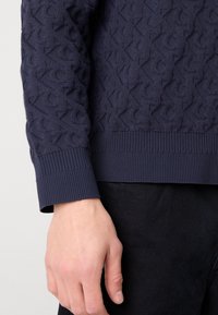 Granatowy sweter wykonany na drutach, z teksturowanym wzorem, ściągaczami na rękawach i miękkim wykończeniem materiału. Zbliżenie na rękaw i dłoń.