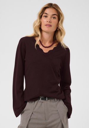 Femme blonde portant un pull à col en V marron foncé et un pantalon à carreaux avec une ceinture noire, accessoirisée d’un collier sombre en perles et de boucles d’oreilles créoles.