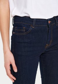 Tumši zili džinsu bikses ar šauru piegriezumu, ar oranžām šuvēm un standarta kabatām. Materiāls izskatās gluds un izturīgs.