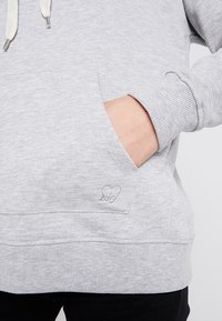 Hand in de voorkant van een lichtgrijze hoodie met ribbelboorden en een klein geborduurd hartdetail nabij de rand van de zak.