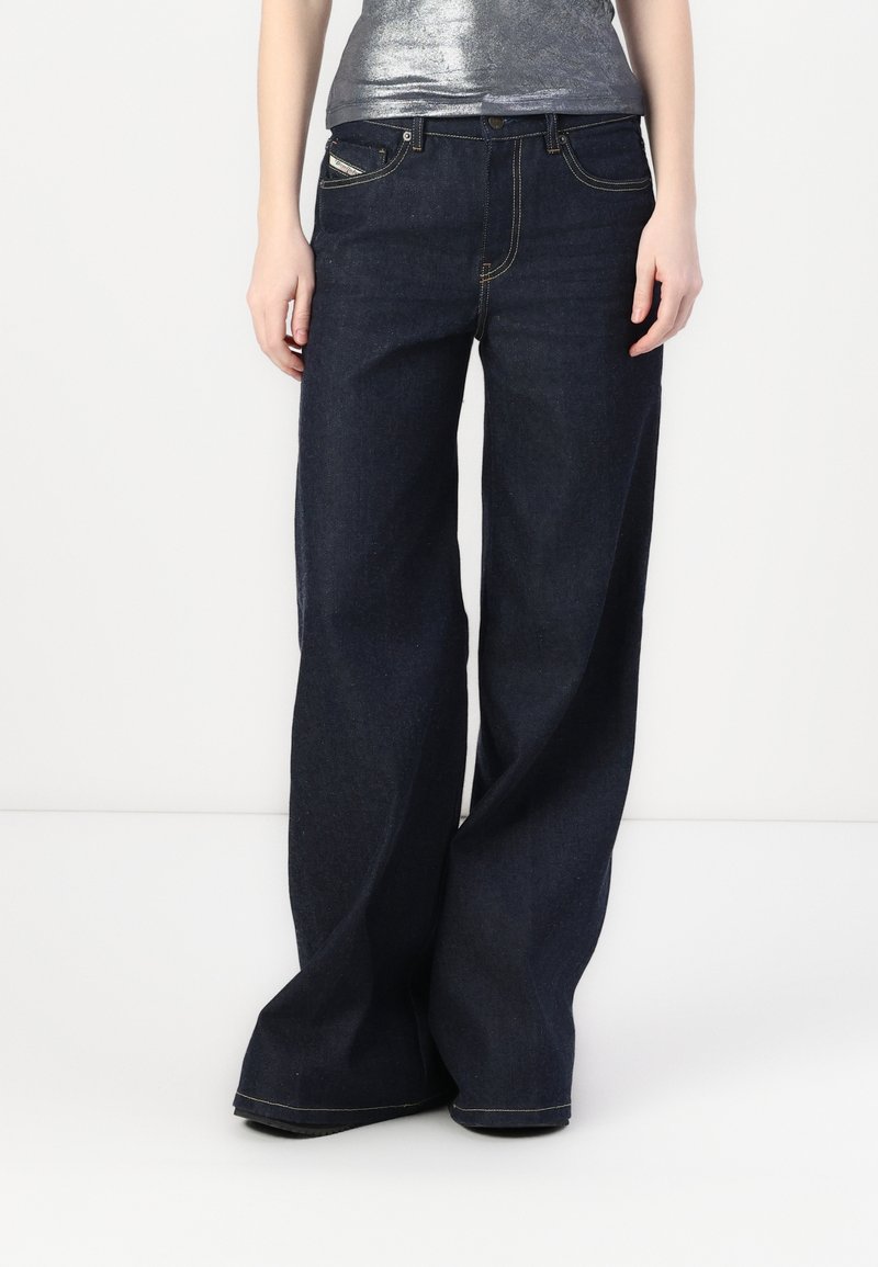Personne portant un jean large bleu foncé et un haut sans manches argent métallique, debout devant un fond blanc uni.