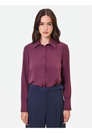 Femme aux cheveux bruns portant un chemisier violet à manches longues boutonné, rentré dans un pantalon taille haute bleu marine avec une ceinture assortie.