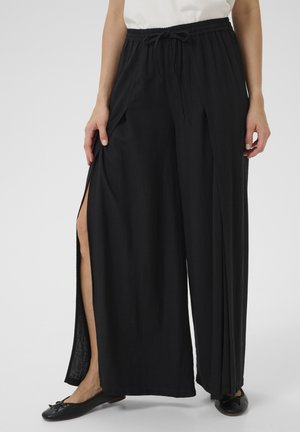 Zwarte wijde broek met een trekkoord in de taille, voorzien van zijslits en een zachte, gestructureerde stof. Gecombineerd met zwarte platte schoenen.