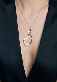 Złoty, abstrakcyjny naszyjnik na delikatnym łańcuszku, noszony przez osobę ubraną w top z głębokim dekoltem w kształcie V. Prosty i elegancki design.