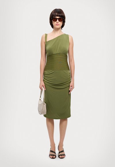 Robe verte olive ajustée, à une épaule, avec un corsage texturé et un détail froncé à la taille. Portée avec des sandales noires et un petit sac léger.