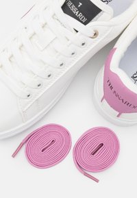 White sneakers with textured upper, pink heel accents, and pink shoelaces displayed alongside. Prominent branding on tongue and heel.