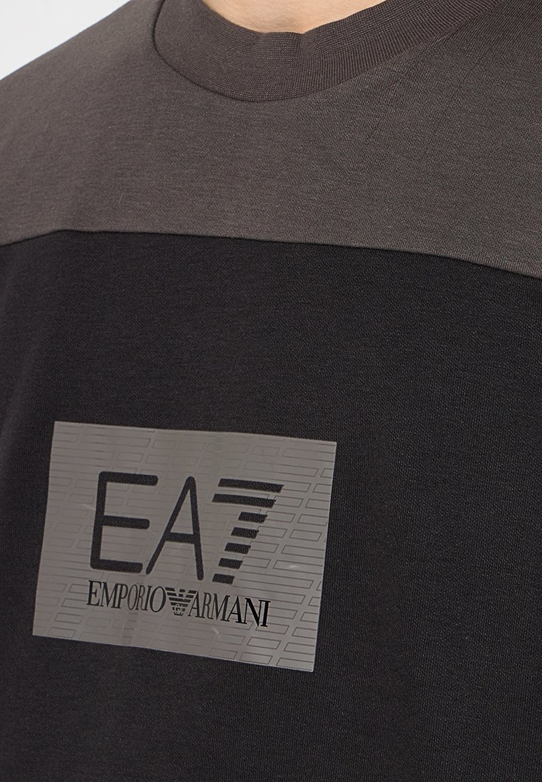 T-shirt nera con un pannello orizzontale grigio, caratterizzata da una toppa logo testurizzata con "EA7" e "EMPORIO ARMANI" in nero. Materiale in cotone.