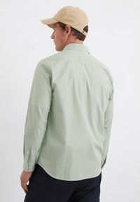 Hellgrünes Langarmhemd mit weißen Streifen; Knopfmanschetten und Kragen. Getragen mit einer beige Kappe, von hinten betrachtet, die die Stoffstruktur zeigt.