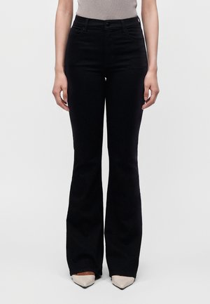 Femme portant un pantalon évasé noir taille haute et des chaussures claires à bout pointu, debout devant un fond clair uni.