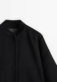 Veste en laine noire avec un col haut, présentant une coupe droite, des manches larges et deux poches avant. Le tissu a une texture lisse.