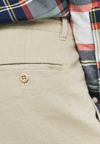 Beige Leinenhosen verfügen über eine Gesäßtasche mit einem Holzknopf und dezentem Logo-Gravur. Das sichtbare Hemd hat ein kariertes Muster in verschiedenen Farben.