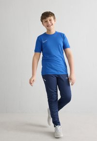 Camiseta deportiva azul de Nike, de manga corta, combinada con pantalones jogger azul marino con detalles blancos. El modelo lleva zapatillas deportivas de color gris claro.
