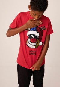 T-shirt rouge avec un graphique de Père Noël en sequins portant des lunettes de soleil et des antennes, texte "JOYEUX NOËL" en bas. Manches courtes, coupe décontractée.