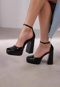 Talons compensés en cuir verni noir avec un large talon carré, bout ouvert et bride ajustable à la cheville. Texture lisse avec finition brillante.