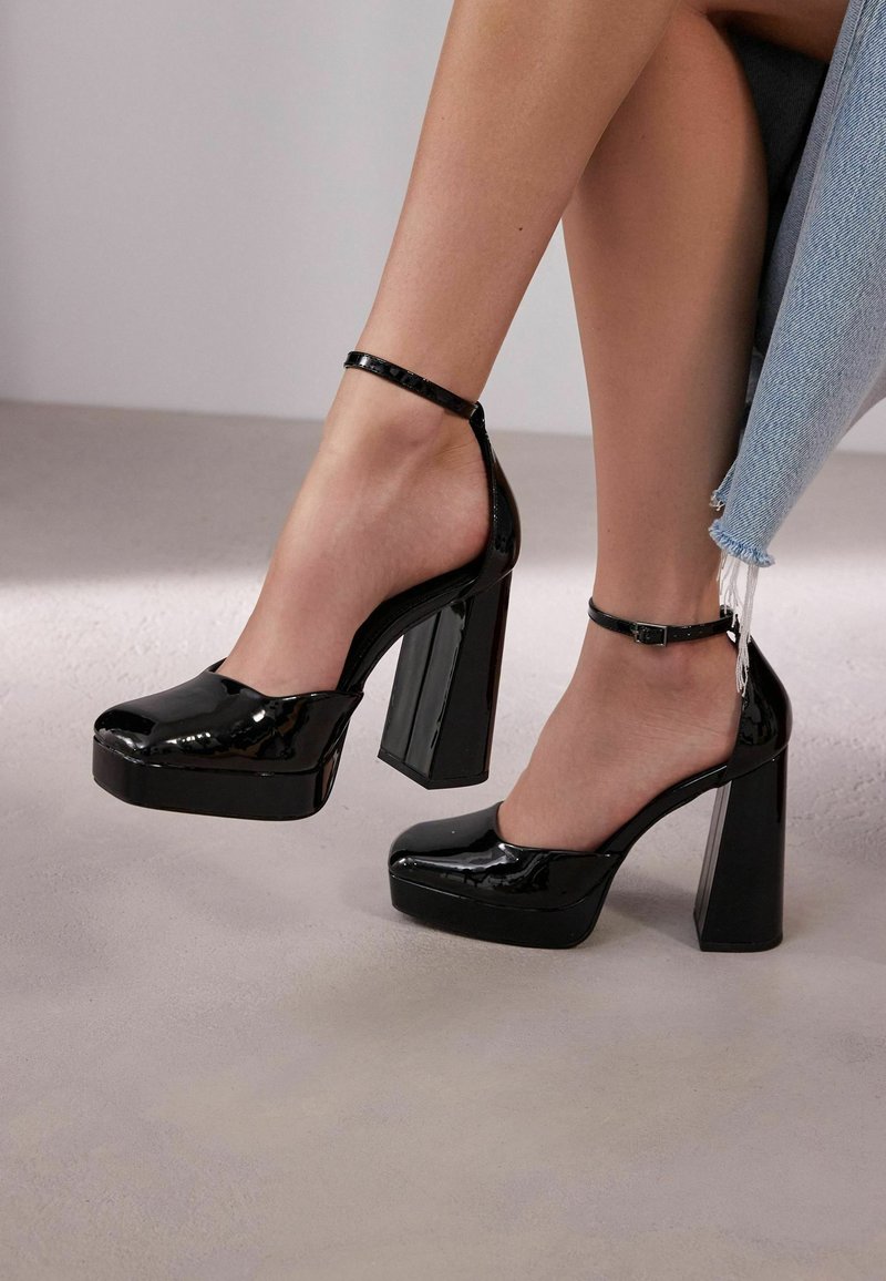 Talons compensés en cuir verni noir avec un large talon carré, bout ouvert et bride ajustable à la cheville. Texture lisse avec finition brillante.