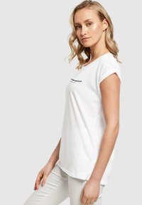 Hvid bomulds t-shirt med opsmøgede ærmer og rund halsudskæring. Har et sort tekstgrafik på forsiden. Glat tekstur og afslappet pasform.