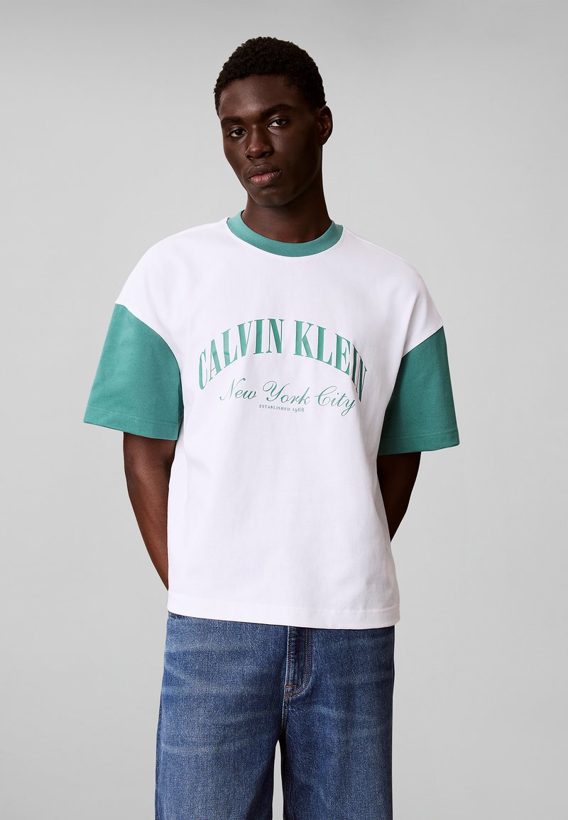 Homme portant un t-shirt blanc et vert Calvin Klein New York City et un jean bleu sur fond gris, debout les mains dans le dos.