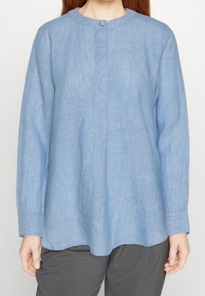 Frau trägt eine hellblaue Leinenbluse mit langen Ärmeln, rundem Ausschnitt und einem einzelnen oberen Knopf, kombiniert mit dunkelgrauen Hosen.