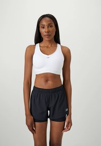 Kvinne som står iført en hvit sports-BH og svarte lagdelte treningsshorts foran en ensfarget bakgrunn.