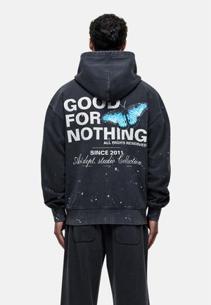 Osoba nosząca czarną bluzę z kapturem z napisem "GOOD FOR NOTHING" i niebieską grafiką motyla na plecach, w połączeniu z czarnymi spodniami.