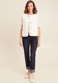 Weißer Fleece-Weste mit goldenen Knöpfen über einem gestreepten Hemd, kombiniert mit dunklen Jeans mit einem gemusterten Saum und hellen Sneakers.