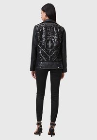 Veste en cuir noir avec un motif complexe de sequins sur le dos, présentant des motifs géométriques. Associée à un pantalon noir ajusté et des chaussures à talons.