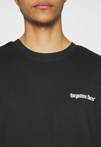 Sort kortærmet T-shirt lavet af bomuld med rund hals; har hvid tekst "glemte ansigtter" i et stiliseret skrifttype på venstre bryst.