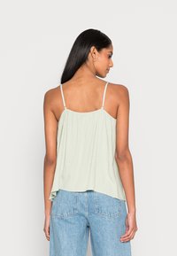Camisola verde-clara, texturizada, com alças finas ajustáveis, apresentando um design fluído que cai ligeiramente mais longo nas costas.