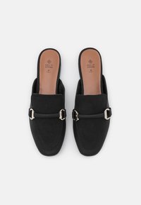 Mules véganes noires à enfiler avec une semelle plate, présentant une finition texturée et un accent de boucle métallique sur le devant. Doublure intérieure douce.