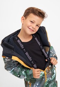 Farverig regnjakke med naturmotiv, udstyret med sort indvendig og lynlås med gule accenter. Sorte "WeeDo" mærkestropper er synlige.