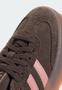 Brown suede sneaker with textured panels, pink stitched stripes on the side, brown laces, and a translucent rubber sole.