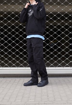 Osoba ubrana w czarne spodnie typu cargo, czarną bluzę z kapturem z białym nadrukiem, jasnoniebieską koszulę pod spodem i czarne tenisówki, stojąca przed metalową bramą.