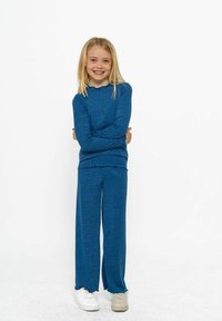 Blå stickad outfit bestående av en långärmad topp med en vågig halsringning och vida byxor med slitsar vid fållen, i kombination med vita skor.