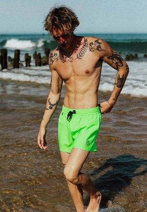 Kale jonge man met meerdere tatoeages, gekleed in felgroene zwembroek, loopt op blote voeten in ondiep oceaanwater met golven en houten palen op de achtergrond.