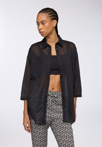 Camicia nera trasparente con bottoni, dal fit rilassato e maniche a tre quarti, indossata sopra un crop top nero e pantaloni con motivo.