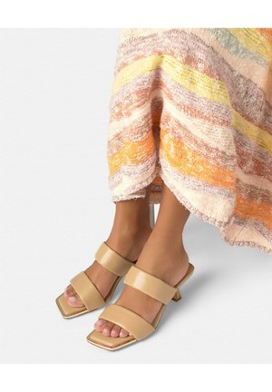 Pies con sandalias de tacón beige de doble tirante y punta cuadrada, junto a una falda multicolor con textura, sobre un fondo blanco.