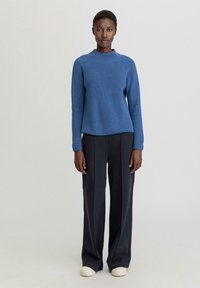 Pull en maille côtelée bleu avec un col rond, porté avec un pantalon taille haute sombre et des baskets claires, se tenant devant un fond neutre.