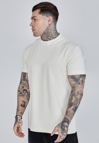 Balta trumpų rankovių marškinėliai su apvaliu iškirpimu, lygia tekstūra ir pritaikytu siluetu. Juos dėvi tatuiruotas asmuo, derinamas su tamsiomis kelnėmis.