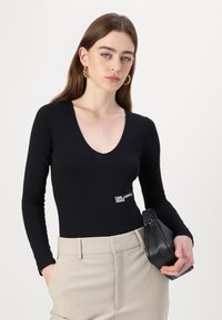 Black long-sleeve bodysuit with a deep V-neck and white "KARL LAGERFELD JEANS" logo. Paired with beige high-waist trousers and a small black bag.