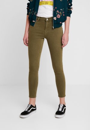 Kvinde iført olivengrønne skinny jeans, sorte og hvide sneakers, en hvid t-shirt med grafik og en mørkegrøn blomstrede lynlåsjakke.