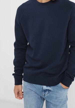 Persoon draagt een marineblauwe trui en lichtblauwe spijkerbroek, met één hand in de zak en de andere ontspannen langs het lichaam.