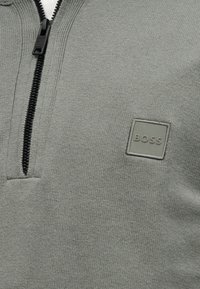 Γκρι πουλόβερ με φερμουάρ και υφή πλεξίματος. Διαθέτει μαύρο φερμουάρ και τετράγωνο ετικέτα με λογότυπο σε ταιριαστή γκρι απόχρωση στο αριστερό στήθος.