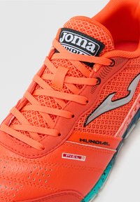 Scarpa atletica arancione con tomaia in mesh, dettagli in pelle sintetica e logo nero. Include lacci piatti e una suola testurizzata.