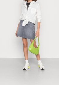 Green glossy handbag with a curved shape, worn on the arm. Paired with a white button-up shirt, gray denim skirt, and sporty shoes.
