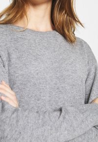 Maglione grigio lavorato a maglia con scollo rotondo, spalle cadenti e maniche lunghe. La texture appare morbida e leggermente pelosa.